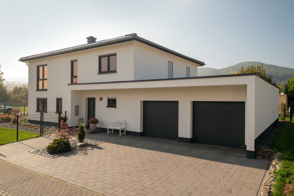 Ein modernes zweistöckiges Haus mit weißen Wänden, schwarzen Fenstern und einem schwarzen Garagentor, umgeben von einem gepflasterten Vorplatz und einem kleinen Garten mit Blumen und Sträuchern, im Hintergrund grüne Hügel und ein klarer Himmel.