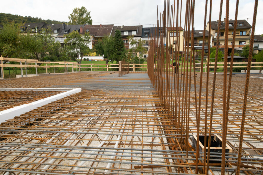 Baustelle mit Stahlbeton-Reinforcement in Baugrund, im Hintergrund sind Häuser mit Gärten und Bäumen zu sehen.