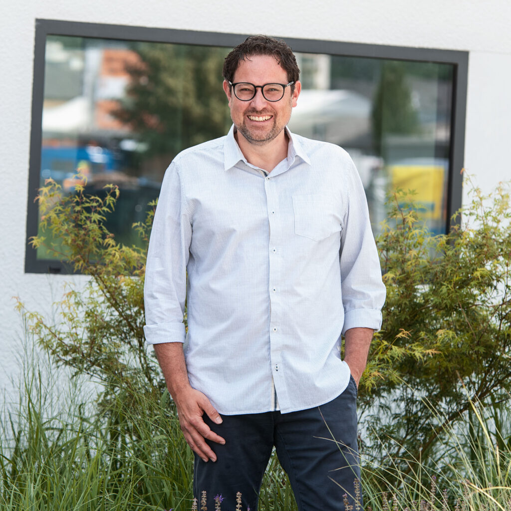 Ein Mann mit Brille, braunen Haaren und Bart steht lächelnd vor einem Gebäude mit einem großen Fenster, umgeben von grünen Sträuchern und Gras.