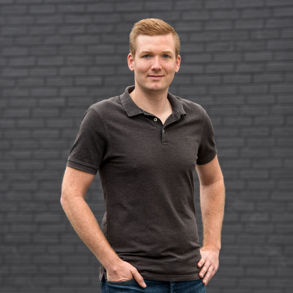 Junger Mann mit roten Haaren, kurzärmligem schwarzen Polohemd und blauen Jeans steht vor einer schwarzen Backsteinmauer und schaut in die Kamera.