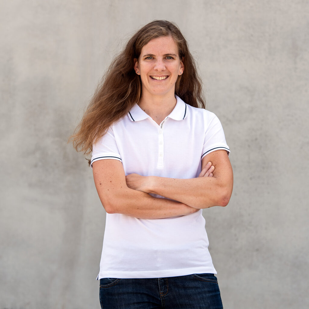 Eine junge Frau mit langem, lockigem braunem Haar, die ein weißes Polo-Shirt trägt und die Arme verschränkt vor einem glatten, hellgrauen Hintergrund stehen lächelt.
