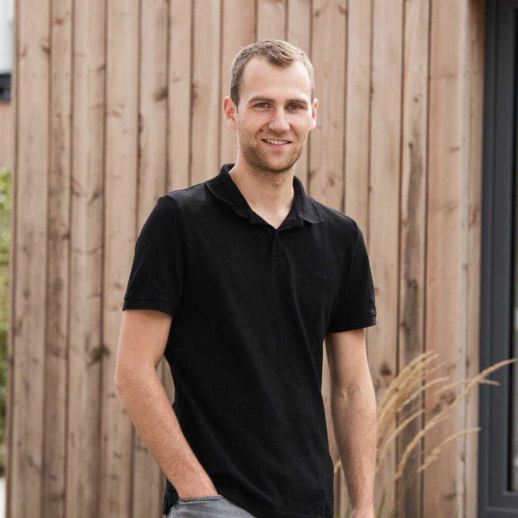 Ein junger Mann mit kurzen braunen Haaren und lightem Bart trägt ein schwarzes Polo-Shirt und steht vor einer Holzwand im Freien, lächelnd in die Kamera blickend.
