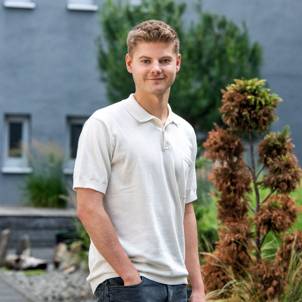 Ein junger Mann mit kurzen, lockigen, hellbraunen Haaren und einem freundlichen Lächeln trägt ein weißes Polo-Shirt und dunkle Jeans vor einer Hauswand mit mehreren Fenstern und einer grünen, verpflanzten Pflanze im Vordergrund.