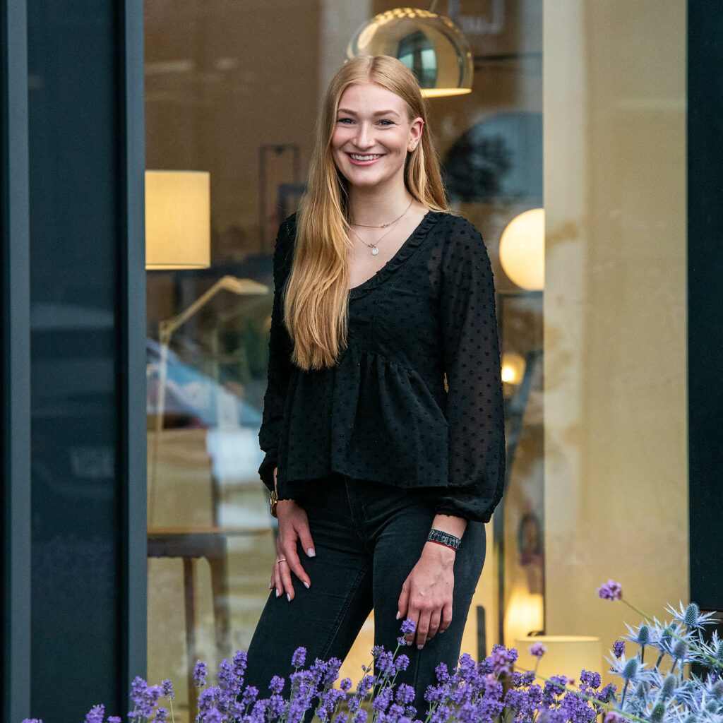 Eine junge Frau mit langen roten Haaren steht vor einem Fenster, trägt eine schwarze Bluse mit Punkten und schwarze Hosen, und lächelt in die Kamera.