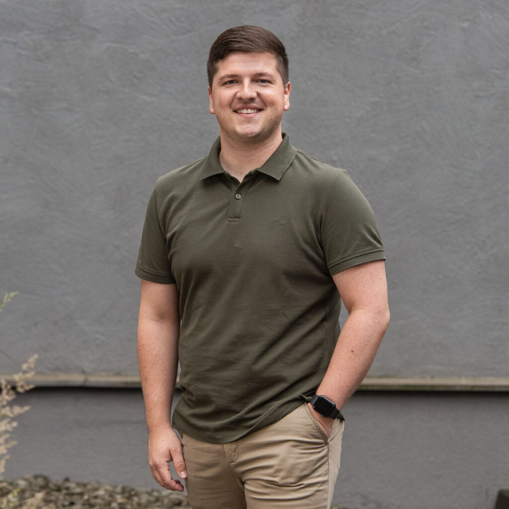 Junger Mann mit kurzen braunen Haaren, lächelnd, trägt ein olivegrünes Polo-Shirt, beige Hosen und eine schwarze Smartwatch, steht vor einer grauen Wand.