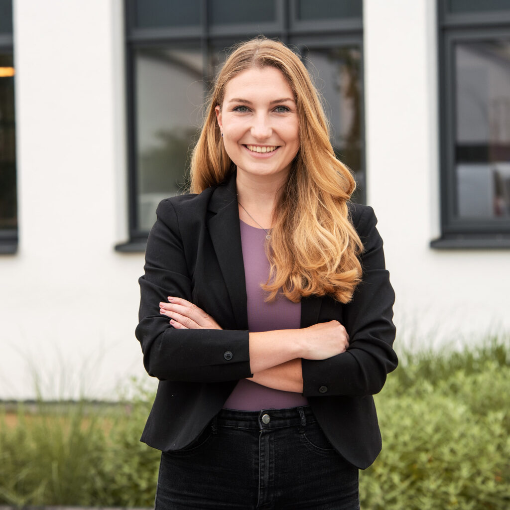 Junge Frau mit langem, gewelltem, blondem Haar, lächelnd und die Arme vor der Brust verschränkt, trägt einen schwarzen Blazer und eine lilafarbene Bluse vor einem modernen Gebäude mit großen Fenstern.