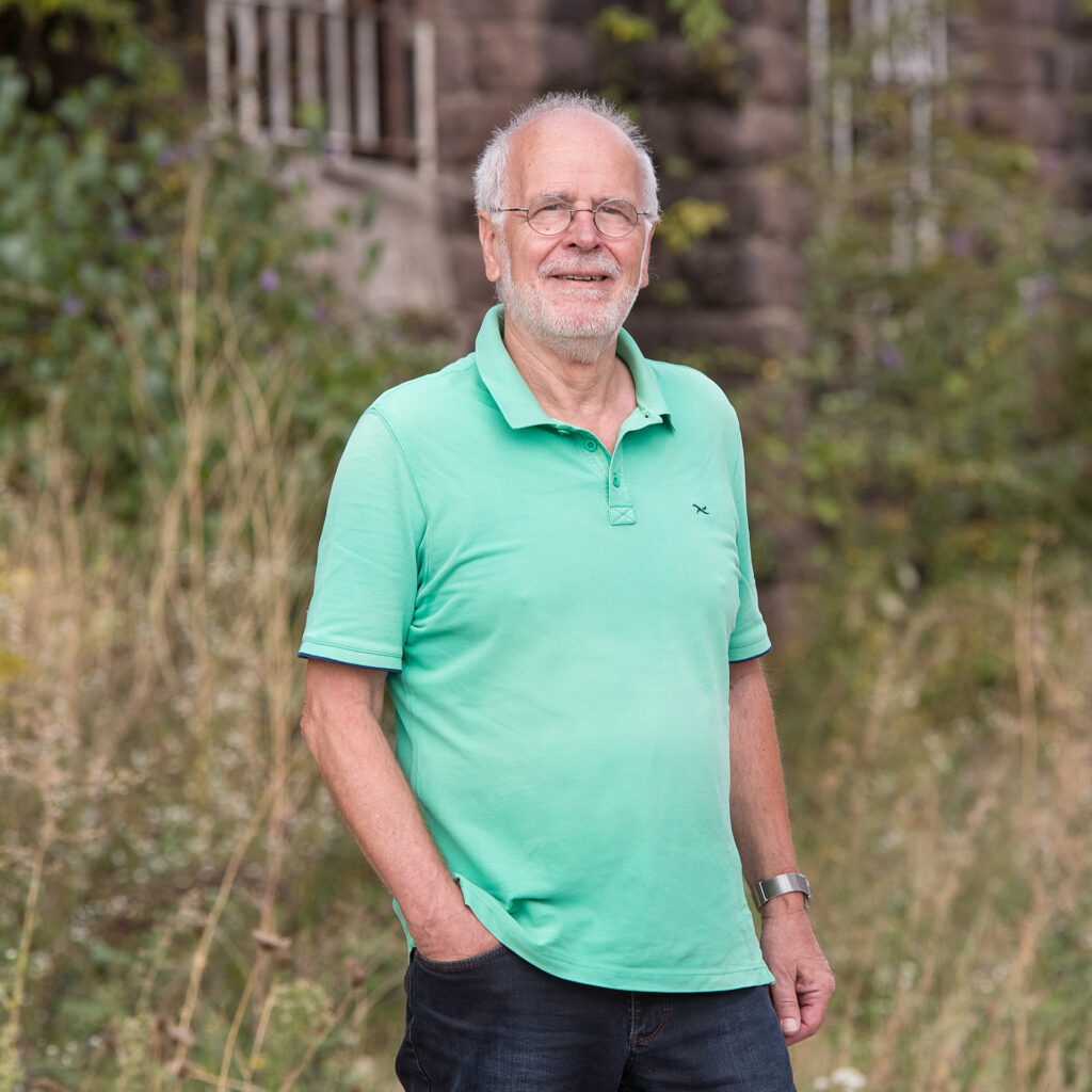 Ein älterer Mann mit weißem Bart und Glasögenden steht im Freien vor einer grünen, bewaldeten Umgebung. Er trägt ein hellgrünes Polo-Shirt und eine Armbanduhr.