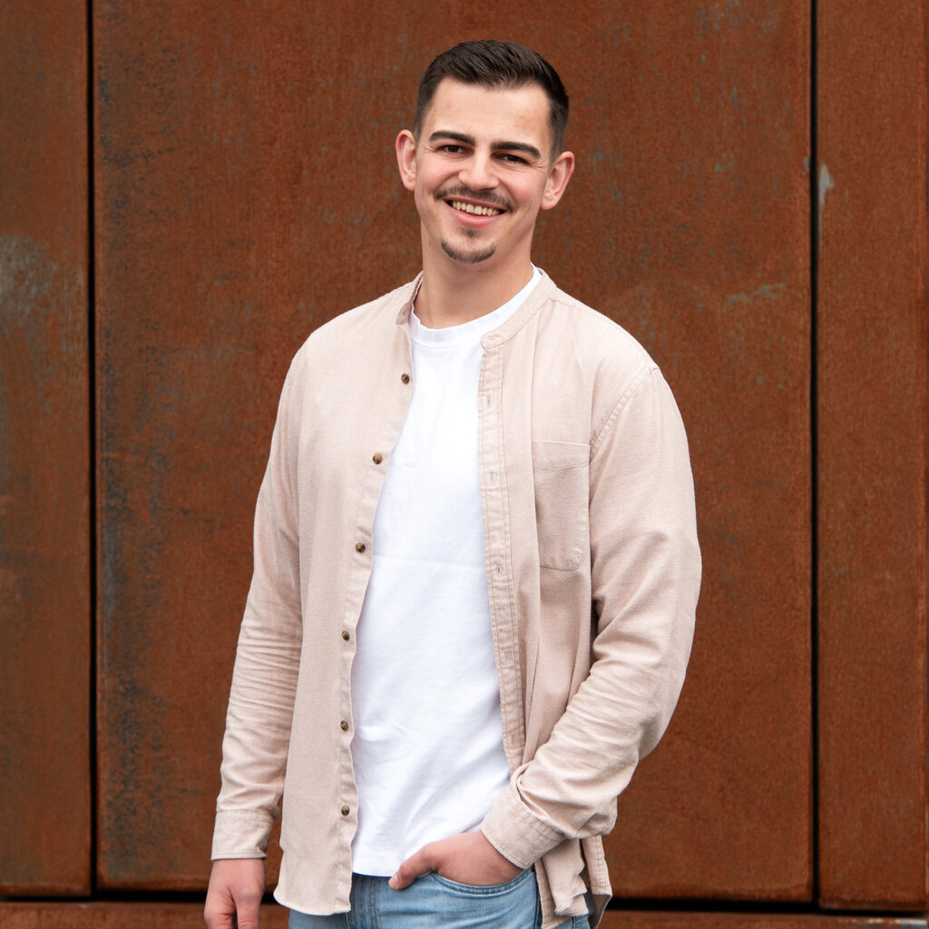 Ein junger Mann mit dunklen Haaren, Bart und Tränensruss trägt ein beige Hemd über einem weißen T-Shirt, steht vor einem rostigen Metallzaun, lächelt und schaut in die Kamera.
