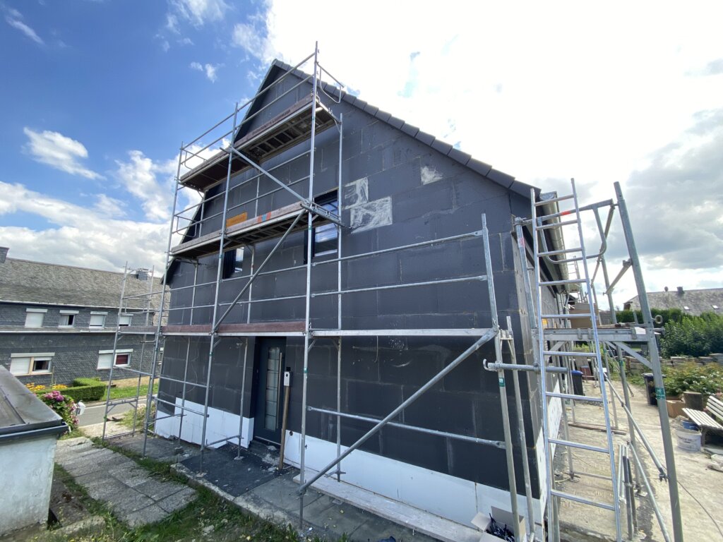 Ein zweistöckiges Haus unter Sanierungsarbeiten, mit schwarzen Dämmplatten an der Fassade, umgeben von Gerüsten und einem blauen Himmel mit Wolken.