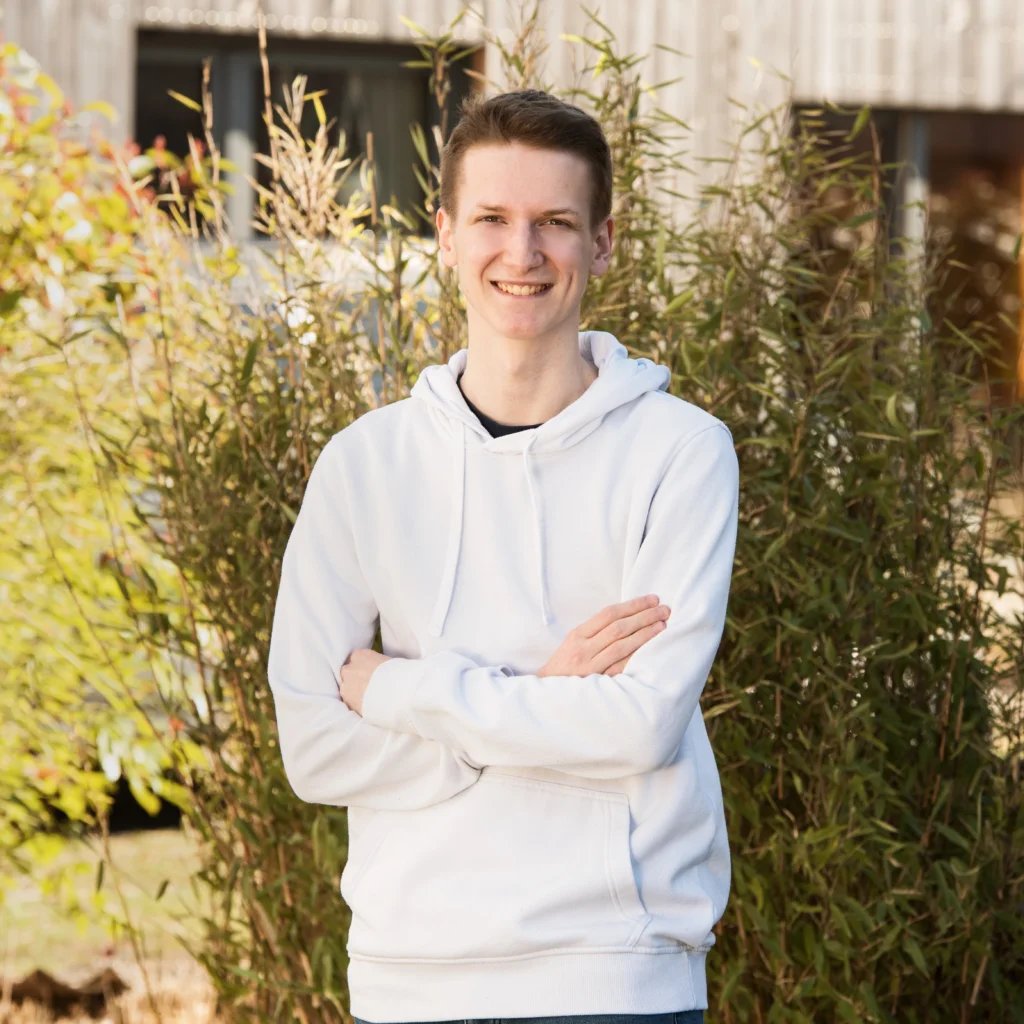 Ein junger Mann mit kurzen braunen Haaren steht im Freien vor einer Hecke mit grünen und gelblichen Blättern, trägt einen weißen Hoodie und lächelt in die Kamera.