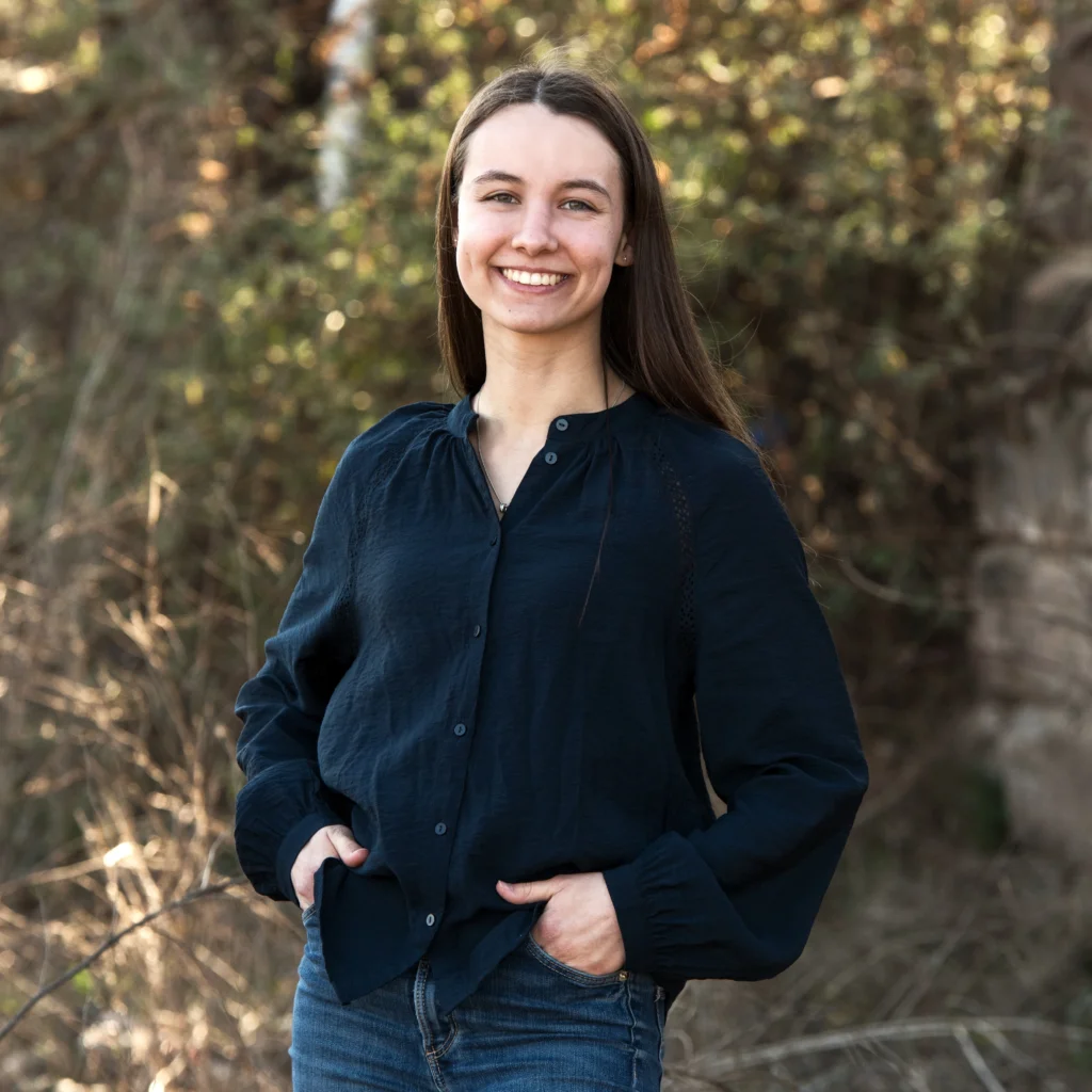 Eine junge Frau mit langen braunen Haaren und einem freundlichen Lächeln, trägt eine dunkelblaue Bluse und Jeans, steht vor einem Hintergrund aus Bäumen und Sträuchern im Freien.