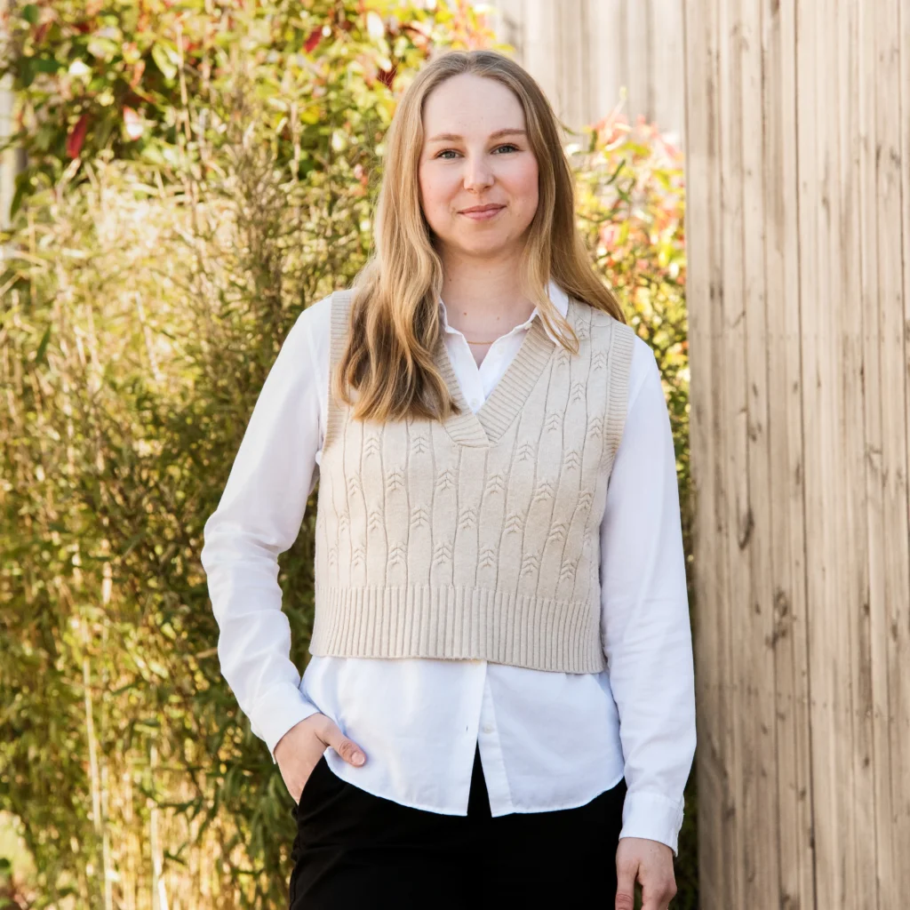Eine junge Frau mit langen, gewellten hellbraunen Haaren steht draußen an einem sonnigen Tag vor einer Holzsicht. Sie trägt eine weiße Bluse und einen beige Walkpulli mit V-Ausschnitt, wobei eine Hand in ihrer Hosentasche steckt.